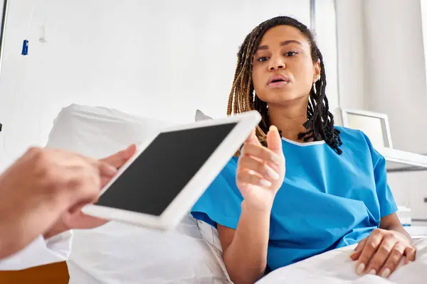 Focus on african american woman in hospital bed looking at her indian doctor taking notes on tablet — Stock Photo