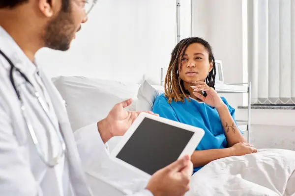 Centrarse en la mujer afroamericana en la cama de hospital mirando a su médico indio tomando notas en la tableta - foto de stock