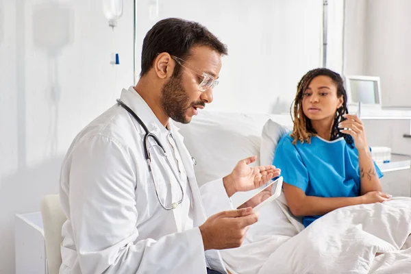 Se centran en médico indio sosteniendo tableta explicando algo a su borrosa paciente afroamericano - foto de stock