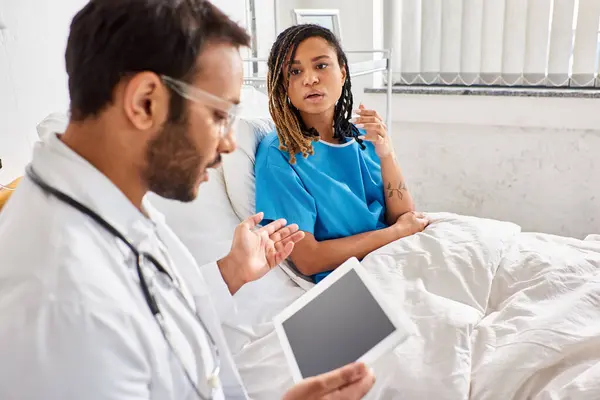 Enfoque en la mujer afroamericana en la cama de hospital mirando al doctor indio borroso con la tableta - foto de stock