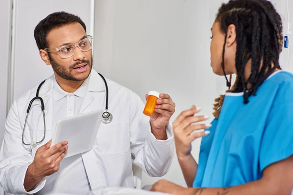 Focus on indian doctor in glasses showing pills to his african american patient holding tablet — Stock Photo