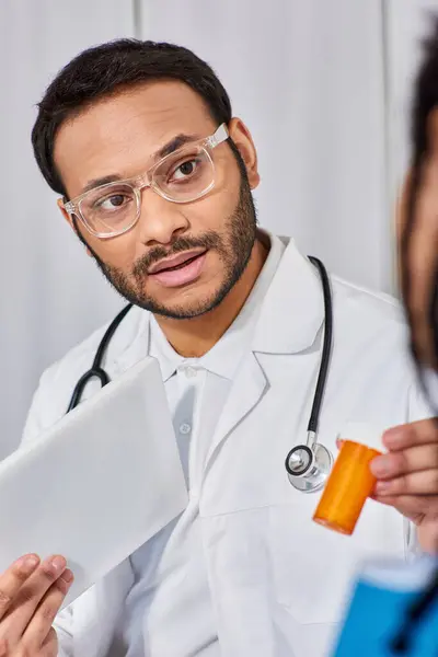 Concentrarsi sul medico indiano in occhiali mostrando pillole al suo paziente afro-americano in possesso di tablet — Foto stock