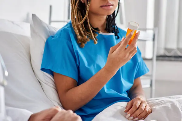 Vista ritagliata di giovane donna afroamericana seduta sul letto d'ospedale e guardando le sue pillole — Foto stock
