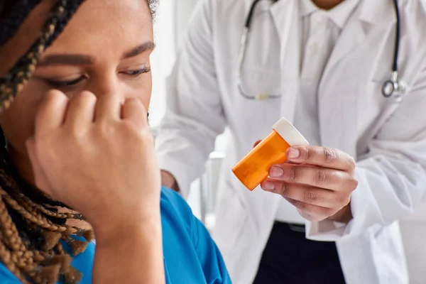 Se centran en las píldoras en manos del médico indio junto a un paciente afroamericano deprimido borroso - foto de stock