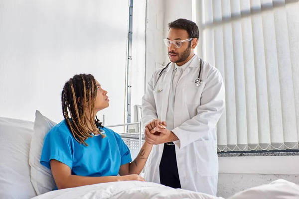 Grave médico indiano confortando seu paciente afro-americano triste em sua ala hospitalar, cuidados de saúde — Fotografia de Stock