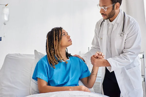 Grave médico indiano confortando seu paciente afro-americano triste em sua ala hospitalar, cuidados de saúde — Fotografia de Stock