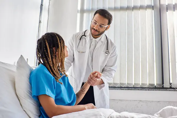 Médico indio alegre consolando a su paciente afroamericano enfermo en su sala de hospital, salud - foto de stock