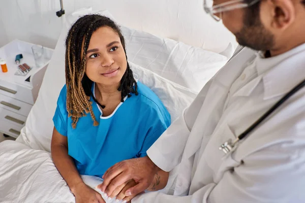 Concentrarsi sulla donna afro-americana allegra in letto d'ospedale tenendo per mano il suo medico indiano offuscata — Foto stock