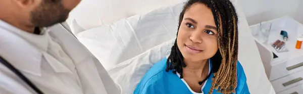 Focus on jolly african american woman in bed looking calmly at her blurred indian doctor, banner — Stock Photo