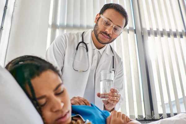 Foco no médico indiano dando copo de água para sua paciente afro-americana doente em sua enfermaria — Fotografia de Stock