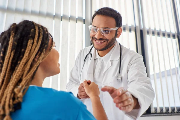 Focus on jolly indian doctor in glasses shaking hand with his blurred african american patient — Stock Photo