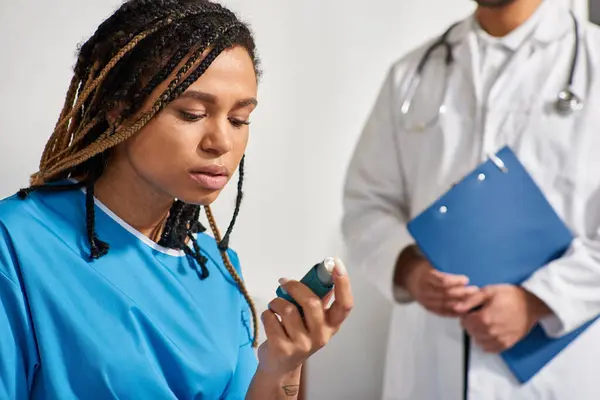Focus on young african american woman using her inhaler with blurred doctor on backdrop, healthcare — Stock Photo
