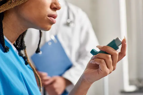 Vista recortada de la joven afroamericana usando su inhalador con médico borroso en el telón de fondo - foto de stock