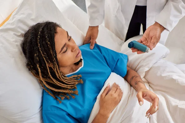 Centrarse en la mujer afroamericana acostada en la cama mientras que el médico indio le enseña a usar inhalador - foto de stock