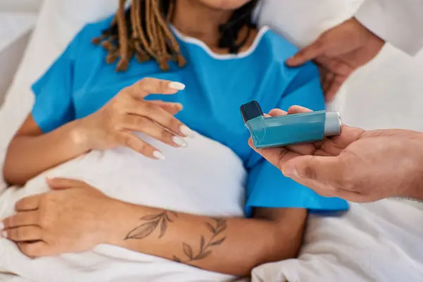 Vista recortada de la mujer afroamericana acostada en la cama mientras que el médico indio le enseña a usar inhalador - foto de stock