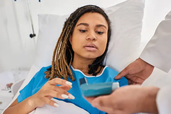 Focus on african american woman lying in bed while indian doctor teach her how to use inhaler — Stock Photo