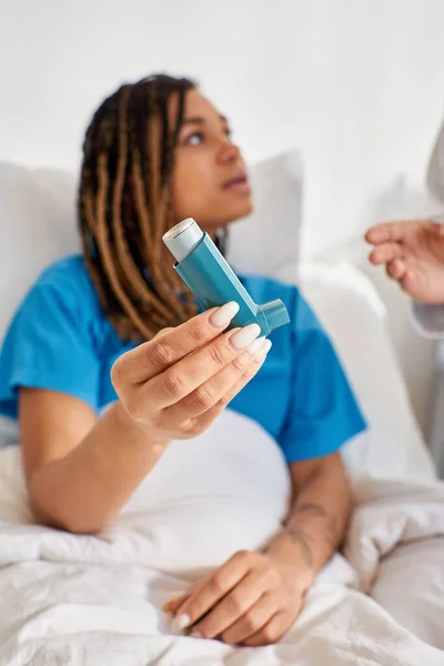Inyección vertical de una joven afroamericana sosteniendo un inhalador de asma y mirando a su médico - foto de stock