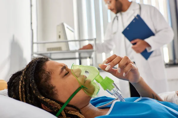 Focus on african american woman lying in bed with oxygen mask with blurred indian doctor on backdrop — Stock Photo