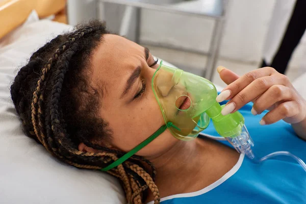 Mujer afroamericana joven enferma acostada en la cama de hospital en su sala con máscara de oxígeno, atención médica - foto de stock
