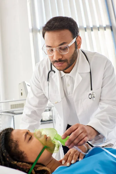 Joven médico indio ayudando a su enfermo africano americano paciente con máscara de oxígeno, atención médica - foto de stock