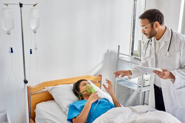 Ill african american woman lying in bed with oxygen mask looking at her indian doctor holding tablet — Stock Photo