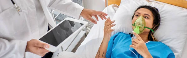 Mujer afroamericana acostada en la cama con máscara de oxígeno mirando al médico indio con tableta, pancarta - foto de stock