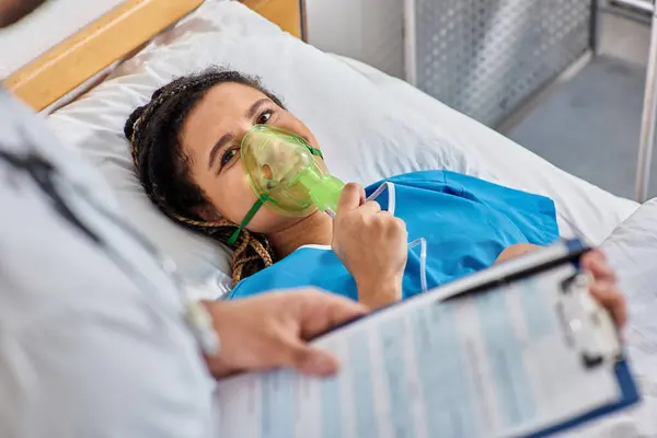 Vista recortada de médico reconfortante mujer afroamericana enferma acostada en la cama de hospital con máscara de oxígeno - foto de stock