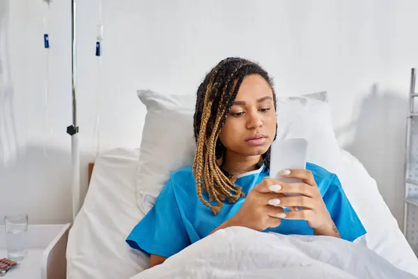 Mujer afroamericana joven acostada en la cama de hospital y mirando su teléfono móvil, atención médica - foto de stock