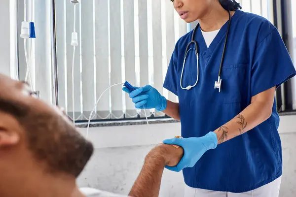 Enfoque en la enfermera afroamericana estableciendo contador de gota junto a su paciente indio desenfocado - foto de stock