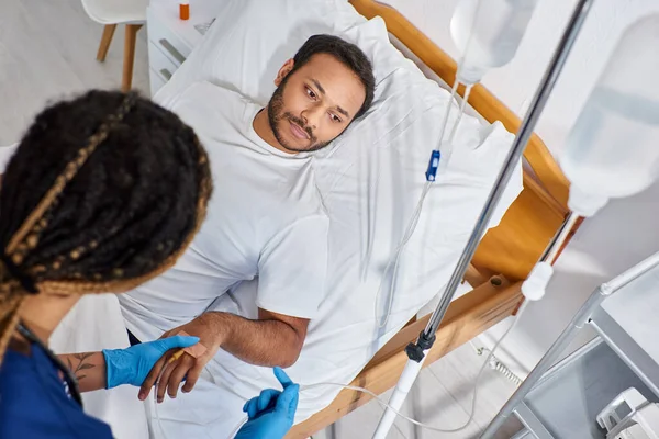 Concentrarsi su uomo indiano sdraiato nel letto d'ospedale, mentre l'infermiera afro-americana impostazione il suo contagocce — Foto stock