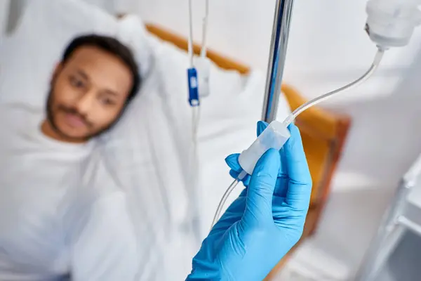 Enfoque en la enfermera afroamericana estableciendo mostrador de gota junto al hombre borroso acostado en la cama de hospital - foto de stock
