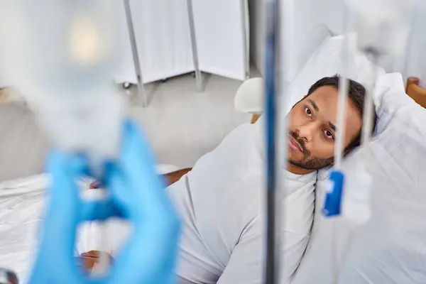 Se centran en el hombre indio enfermo acostado en la cama del hospital en la sala y mirando a la cámara a través de mostradores de gota - foto de stock