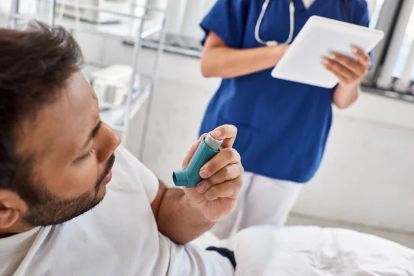 Vista recortada del hombre indio que usa inhalador de asma con enfermera borrosa en el telón de fondo, atención médica - foto de stock