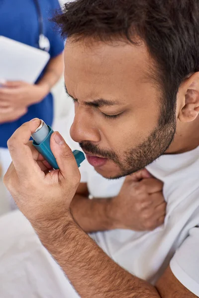 Cropped view of indian man using asthma inhaler with blurred nurse on backdrop, healthcare — Stock Photo