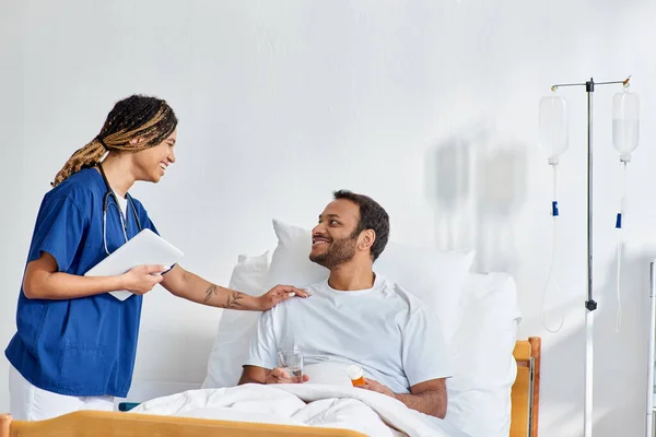 Joyeux afro-américain infirmière souriant joyeusement à son patient indien qui se trouve dans le lit d'hôpital — Photo de stock