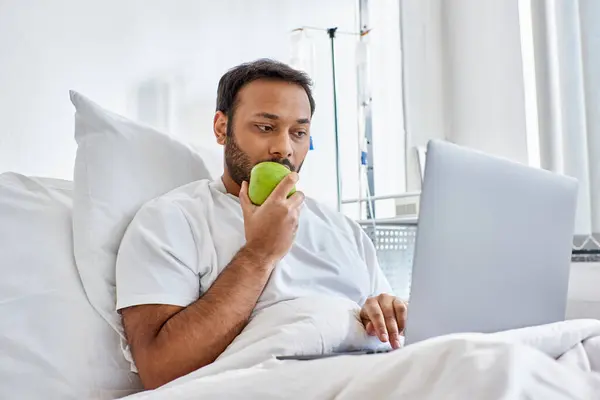 Jeune homme indien ayant un appel vidéo et tenant pomme verte tout couché dans le lit d'hôpital, soins de santé — Photo de stock