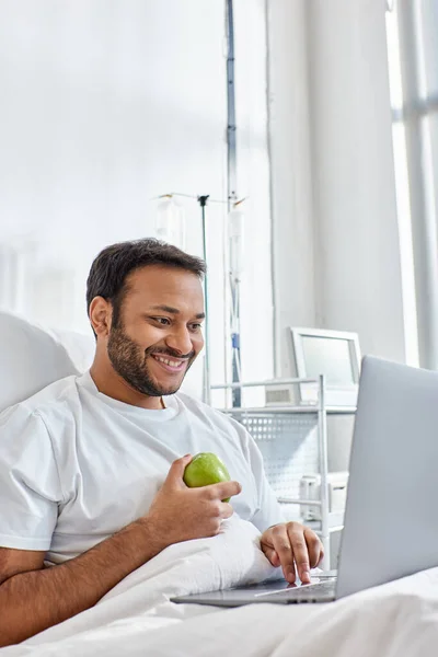 Joven indio hombre teniendo videollamada y sosteniendo manzana verde mientras está acostado en la cama del hospital, atención médica - foto de stock