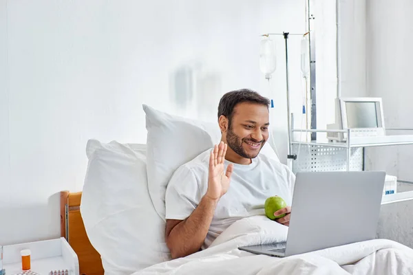 Jovem indiana homem ter vídeo chamada e segurando maçã verde enquanto deitado no hospital cama, cuidados de saúde — Fotografia de Stock