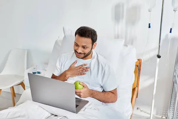 Giovane indiano che fa videochiamate e tiene in mano la mela verde mentre è sdraiato nel letto d'ospedale, assistenza sanitaria — Foto stock