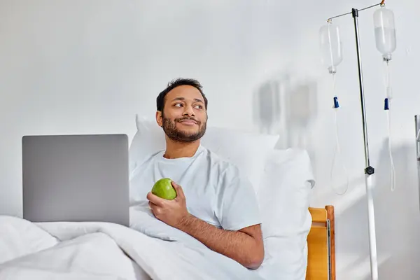 Alegre indiana homem deitado no hospital cama segurando seu laptop e maçã verde e olhando para longe — Fotografia de Stock