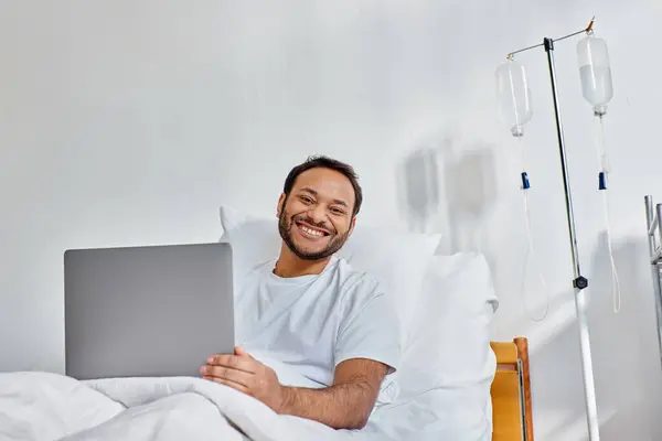 Joven hombre indio alegre sonriendo a la cámara mientras trabaja en el ordenador portátil en la cama del hospital, atención médica - foto de stock