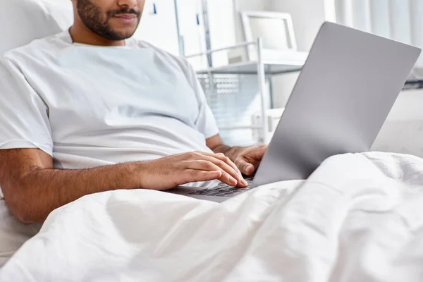 Vista cortada do paciente indiano deitado na cama do hospital em sua ala olhando para seu laptop, cuidados de saúde — Fotografia de Stock