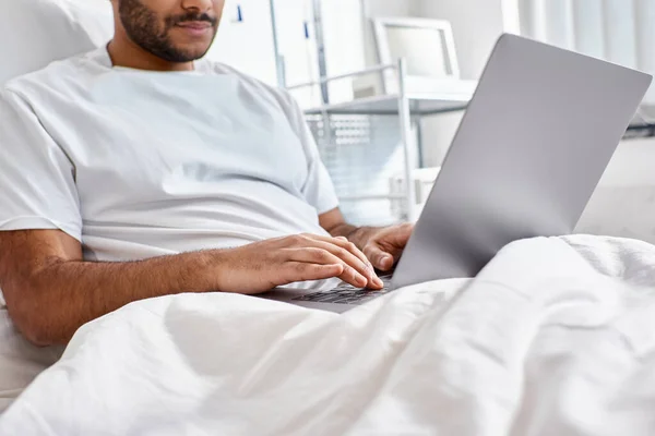 Cropped view of indian patient lying in hospital bed in his ward looking at his laptop, healthcare — Stock Photo
