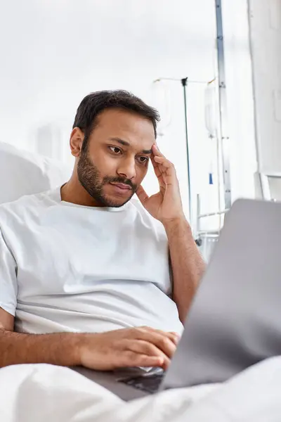Paciente indiano concentrado deitado na cama do hospital em sua ala olhando para seu laptop, cuidados de saúde — Fotografia de Stock
