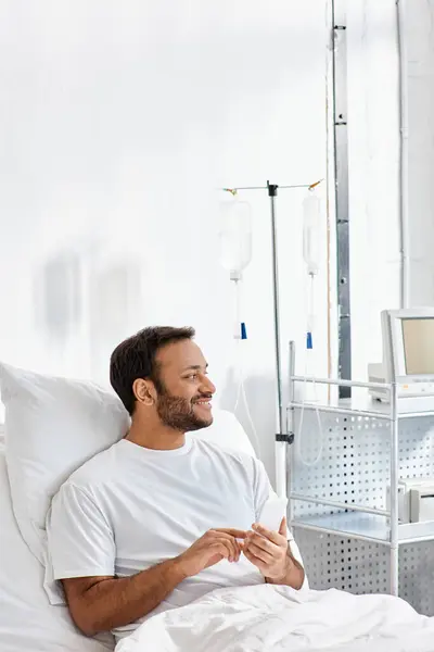 Homem indiano alegre deitado na cama do hospital com telefone nas mãos e sorrindo alegremente, cuidados de saúde — Fotografia de Stock