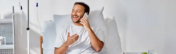 Alegre joven indio hombre hablando alegremente por teléfono mientras está acostado en la cama en la sala de hospital, pancarta - foto de stock