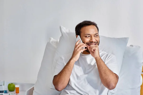 Веселый молодой индийский мужчина весело разговаривает по телефону, лежа в постели в больничном отделении, здравоохранения — стоковое фото