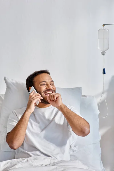 Jovem índio alegre falando alegremente por telefone enquanto estava deitado na cama na enfermaria do hospital, cuidados de saúde — Fotografia de Stock