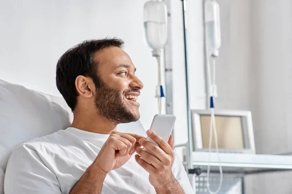 Homem indiano alegre deitado na cama do hospital com telefone nas mãos e sorrindo alegremente, cuidados de saúde — Fotografia de Stock