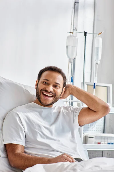 Joyeux jeune homme indien allongé dans le lit d'hôpital et souriant joyeusement à la caméra, soins de santé — Photo de stock